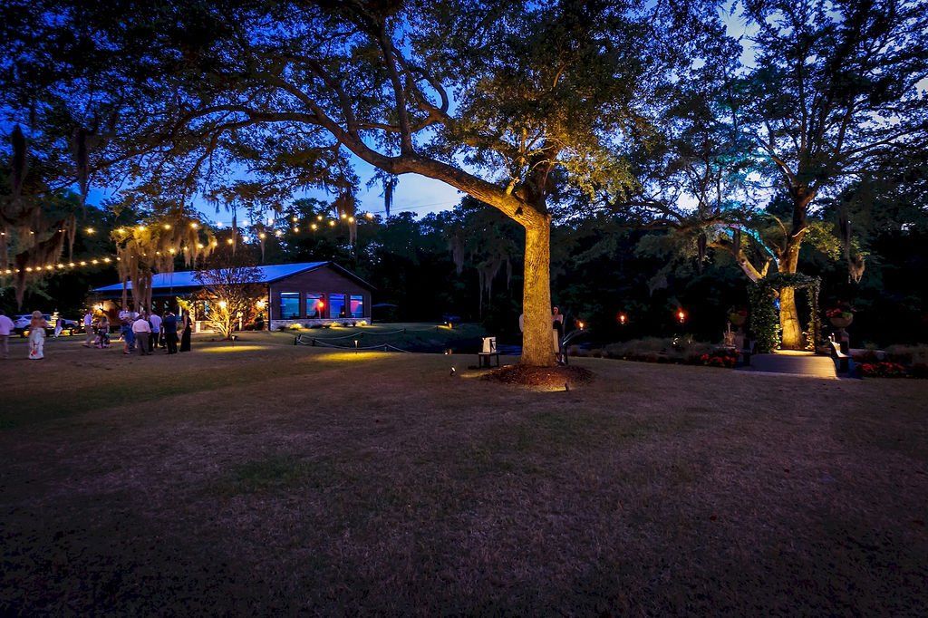 Twilight outdoor park with large trees, lit pavilion, and string lights over a grassy lawn