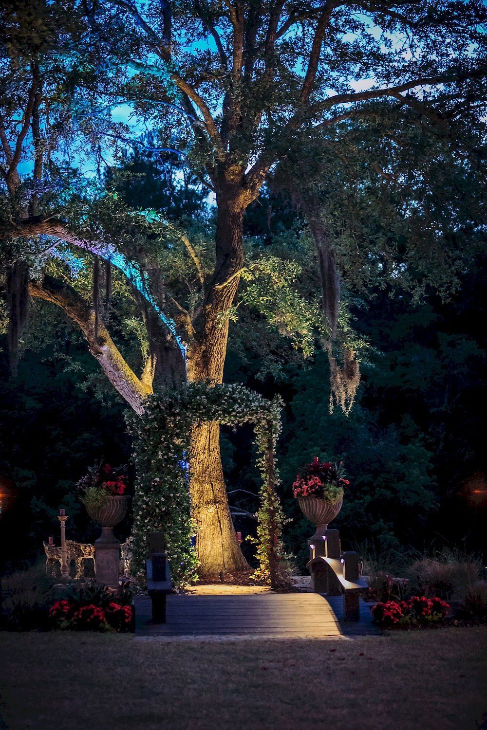 A lit garden staircase under tall trees, framed by flowers at dusk.