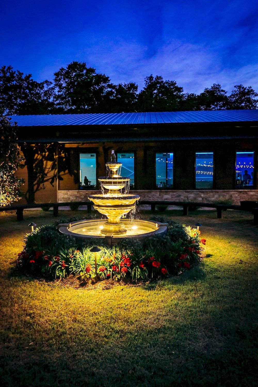 Lit fountain glowing in a flowerbed outside a building at dusk beneath a deep blue sky