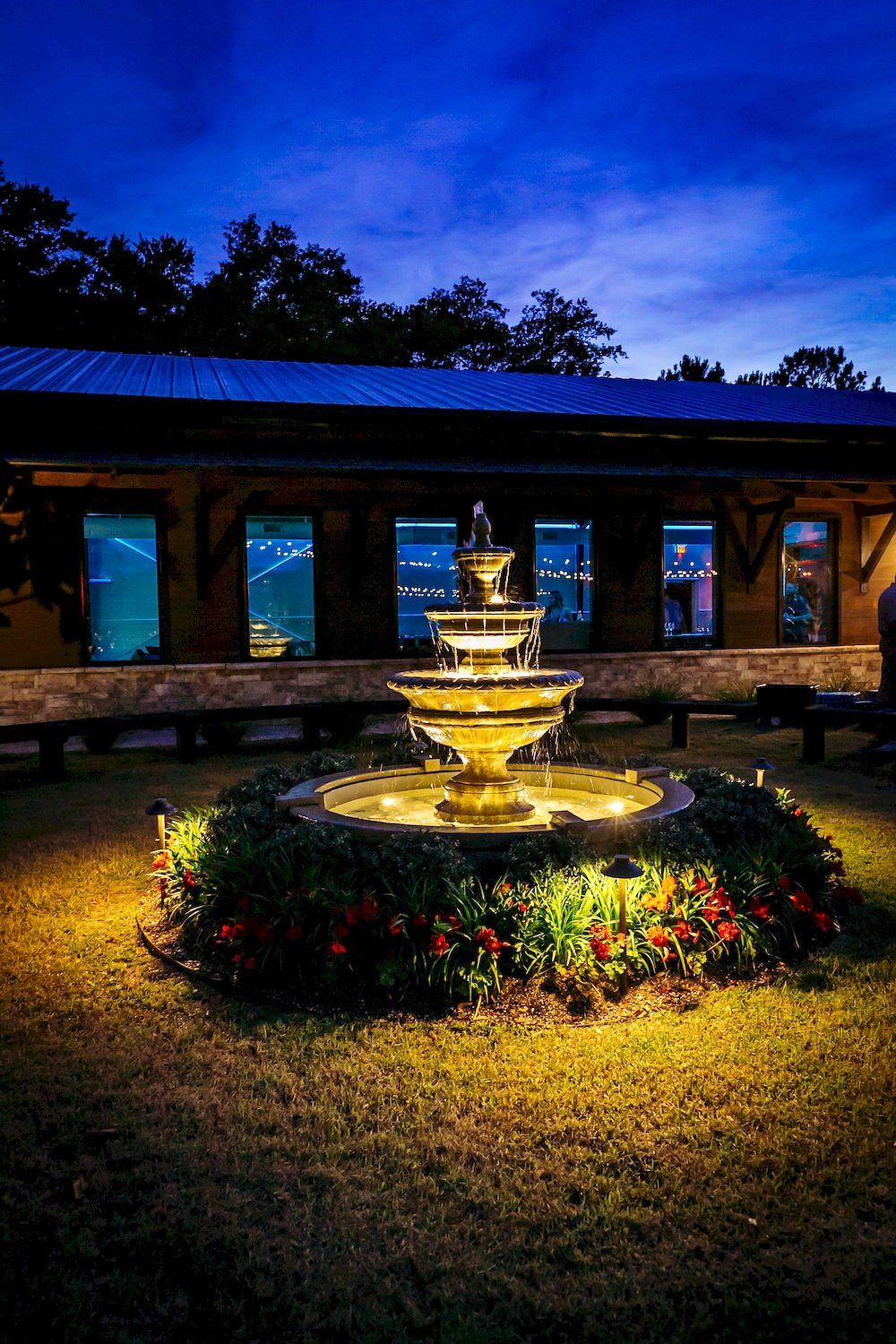 Illuminated fountain in a courtyard at dusk, with flowers and a building silhouetted against a blue sky