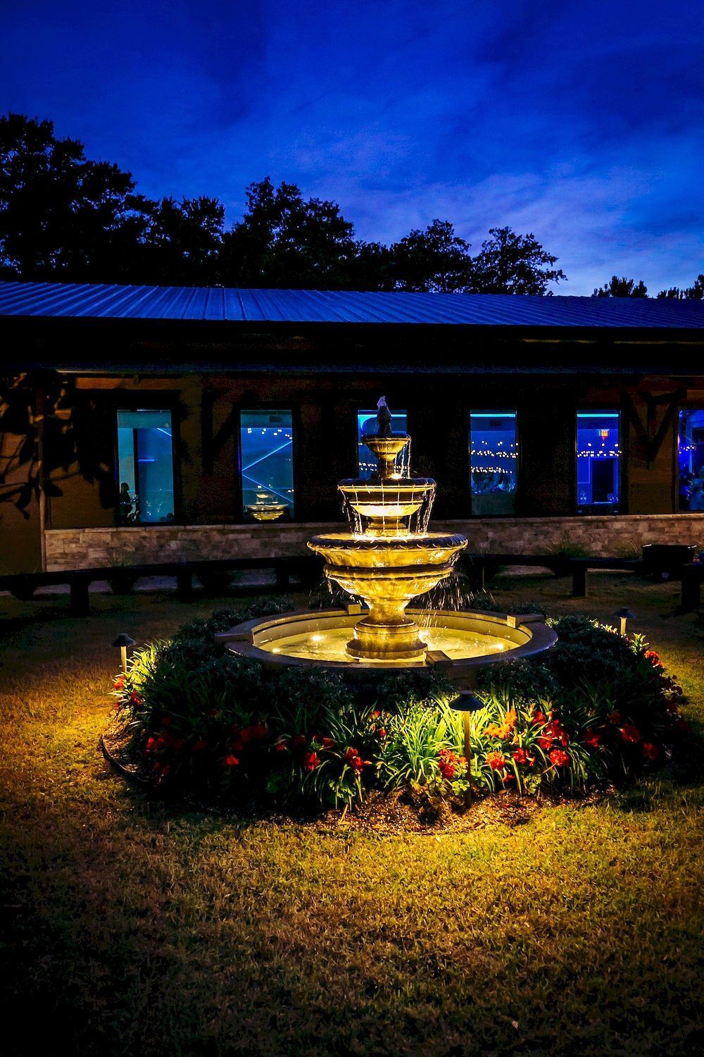 Illuminated fountain in a garden courtyard at night, with blue-lit windows and dark trees in the background