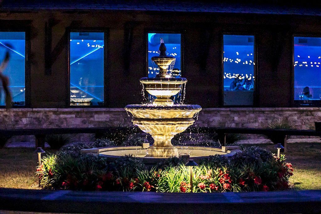 Illuminated tiered fountain at night in a landscaped courtyard with flowers and blue-lit windows