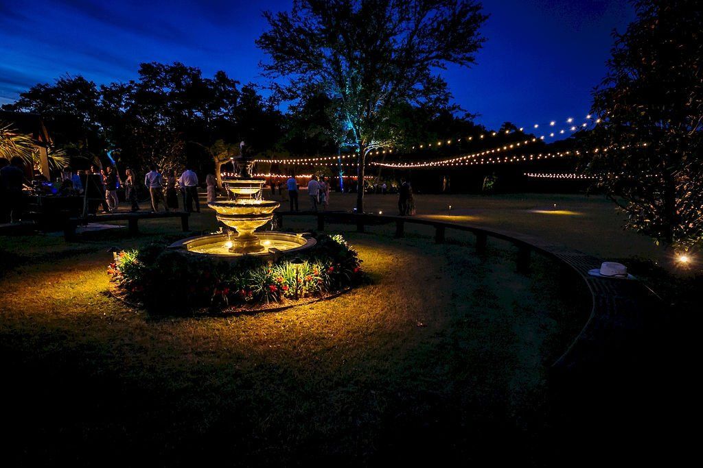 Illuminated fountain in a park at night, with string lights and people walking along the path