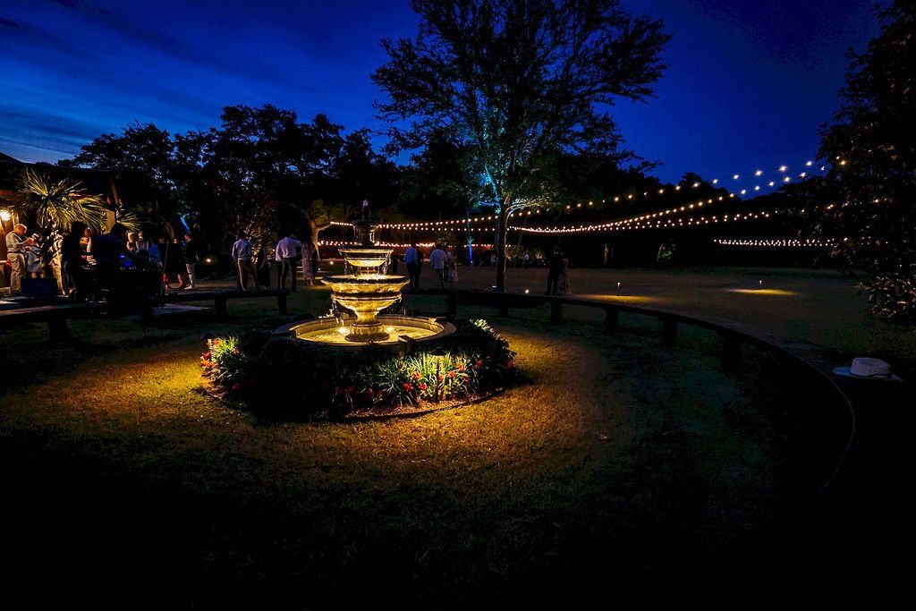 Nighttime garden with a lit fountain, string lights, and silhouetted trees along a walkway