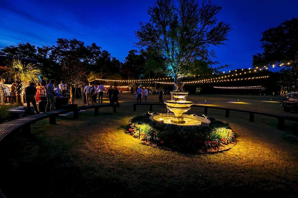 Nighttime garden with a lit fountain, string lights, and people gathered along a path