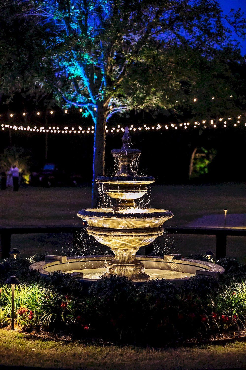 Illuminated tiered fountain in a garden at night with string lights and colorful trees beyond.