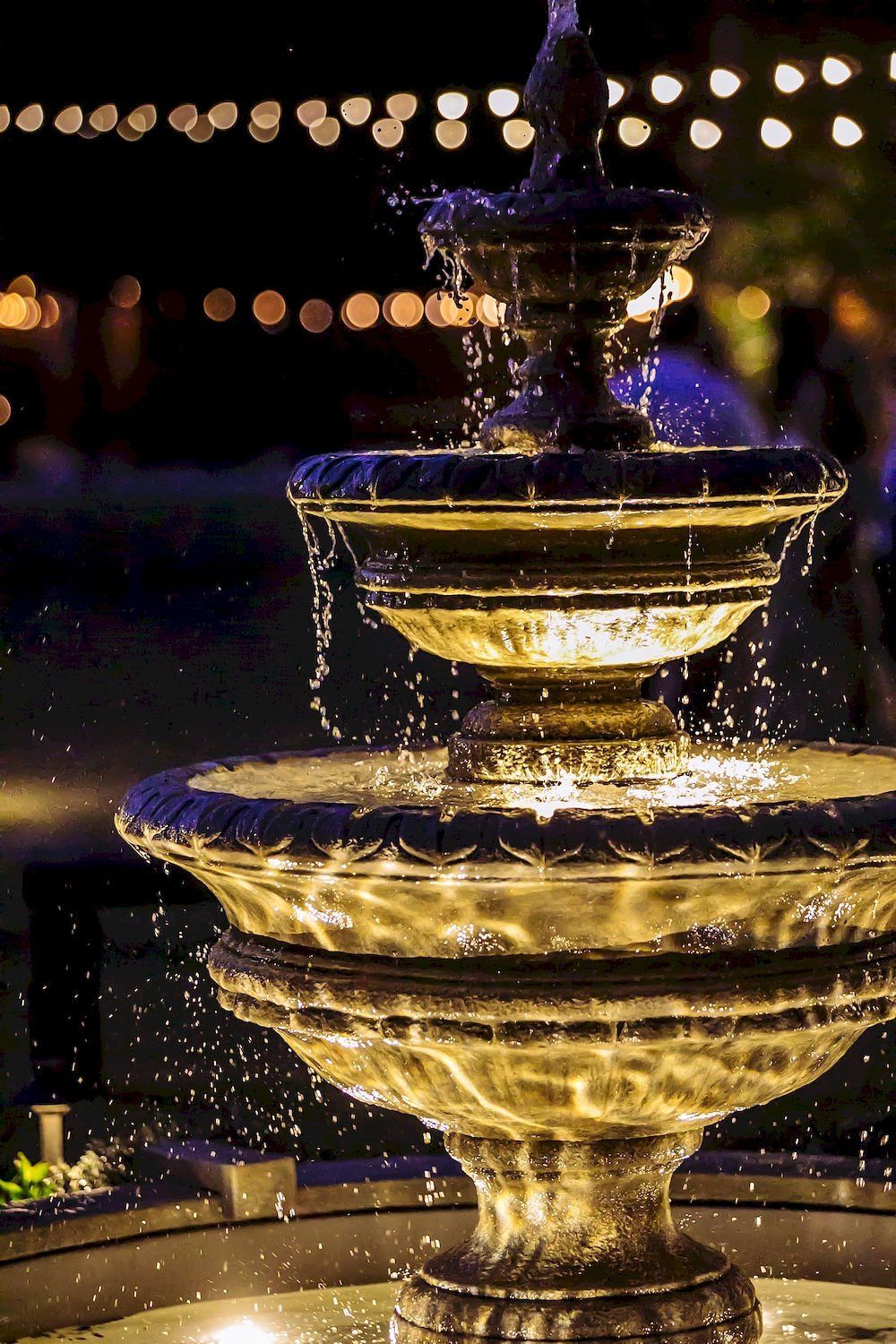 Illuminated tiered fountain with water cascading at night, with warm lights in the background