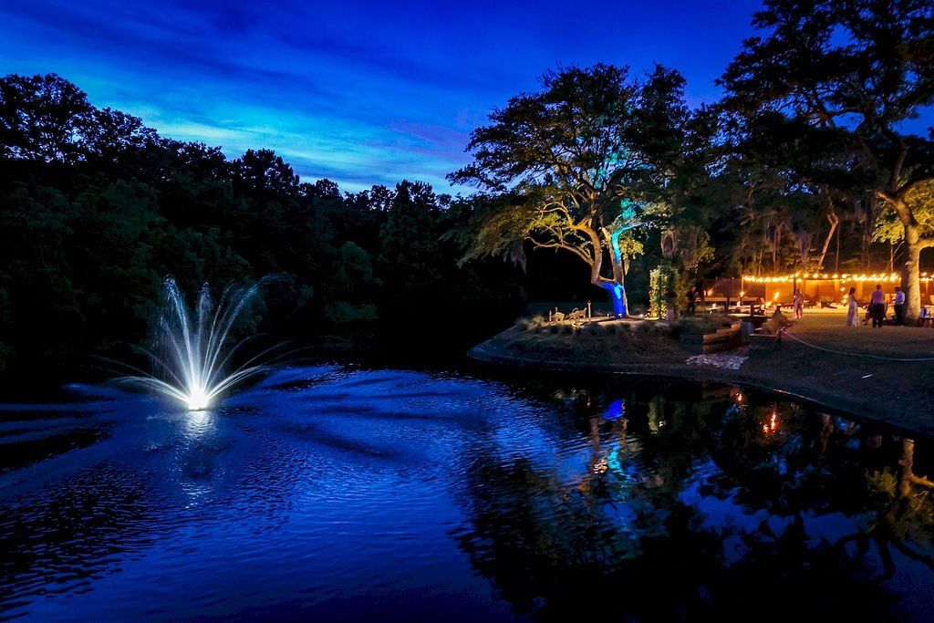 Lit fountain on a pond at dusk, with glowing trees and lights reflecting on the water.