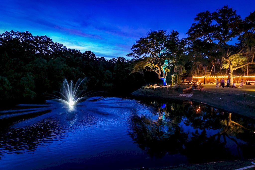 Lit park pond at dusk with a fountain, trees, and blue water reflecting lights and sky