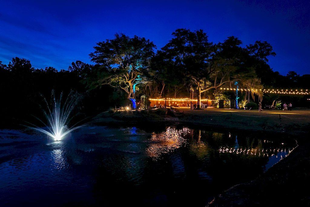 Nighttime park pond with a lit fountain, glowing trees, and reflections on the water