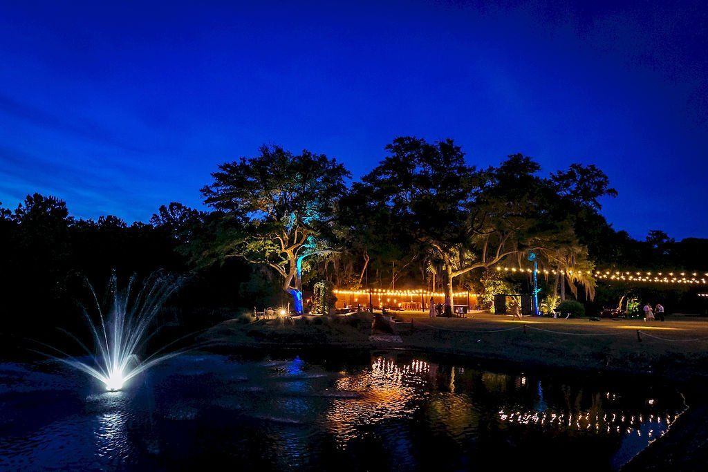Nighttime park pond with fountain, glowing lights, trees, and blue sky reflections