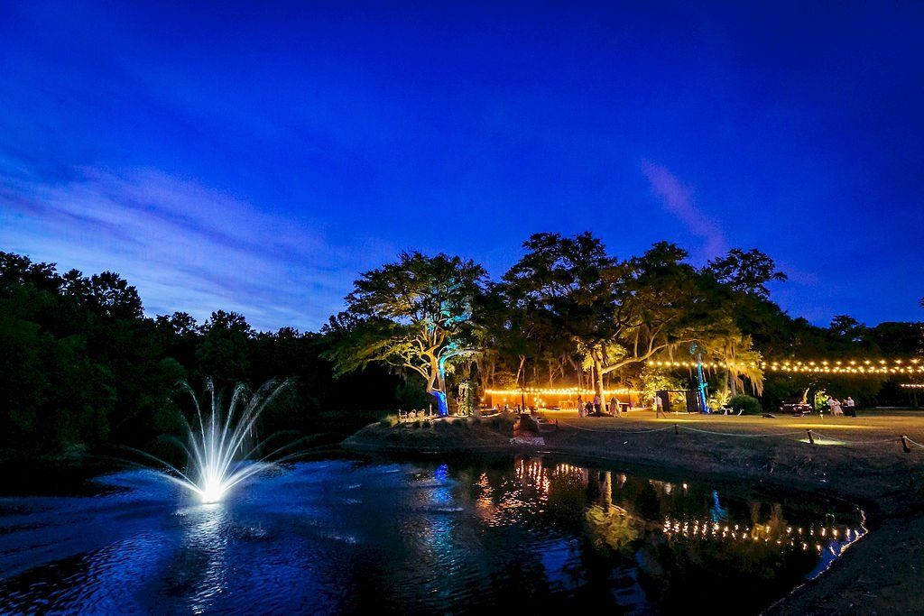 Nighttime park pond with lit fountain, glowing trees, and reflections under a deep blue sky