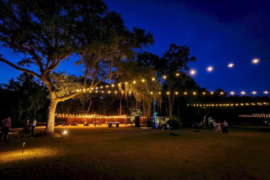 String lights over a grassy park at dusk, with trees and a small seated group in the distance