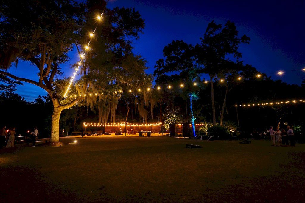Nighttime outdoor gathering with string lights, trees, and a lit stage in a park