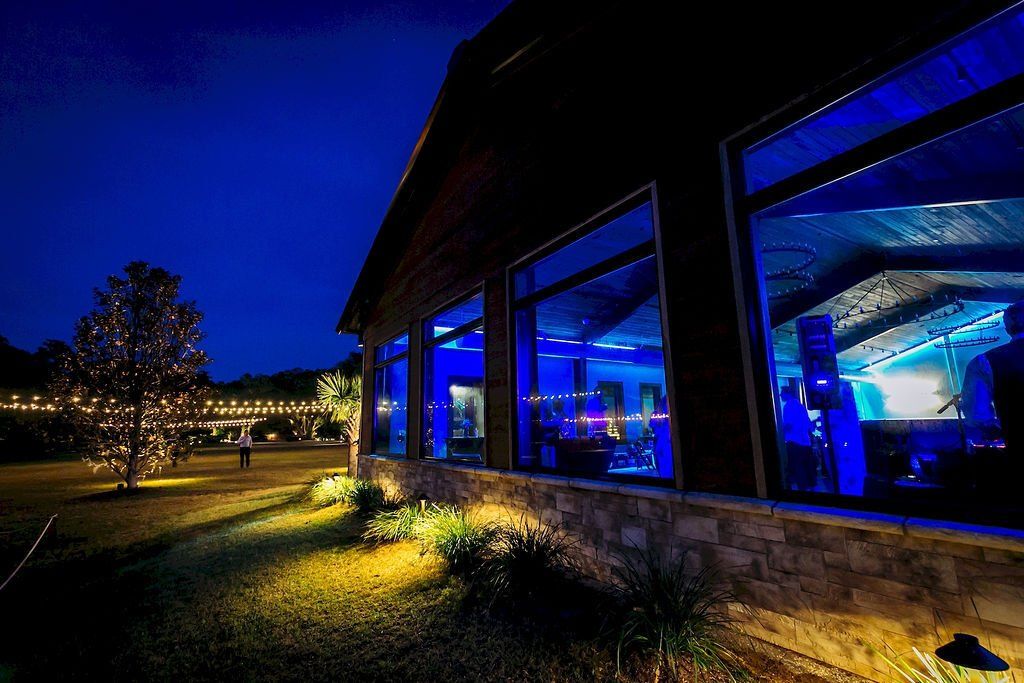 Blue-lit modern building at night with large windows, lit lawn, and a tree in the foreground