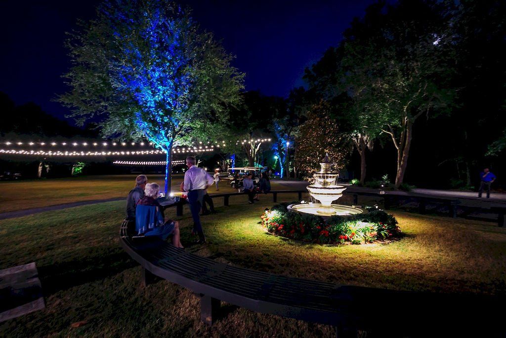 Nighttime park with lit trees, people seated on benches, and a flowerbed around a fountain.