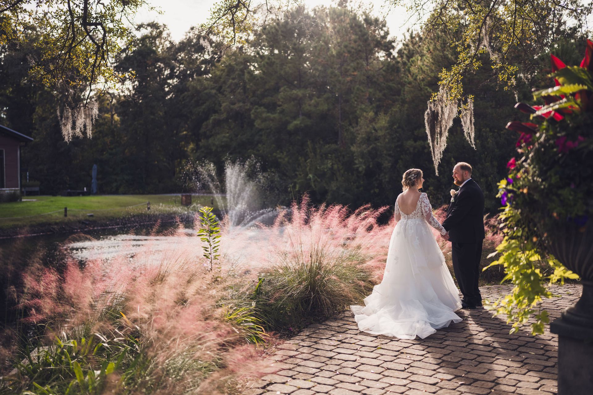 Bride and groom stand on a garden path beside pink grasses and a fountain in soft evening light