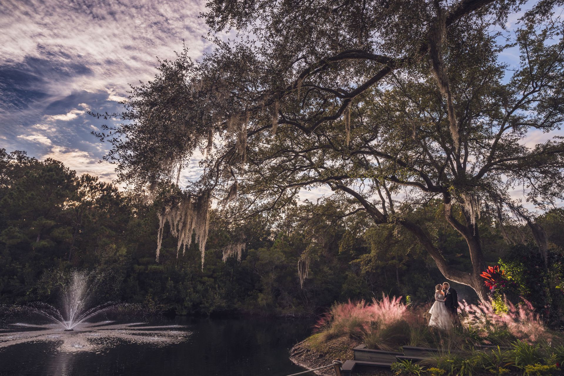 Tree-lined pond at dusk with a fountain, pink flowers, and a colorful sky