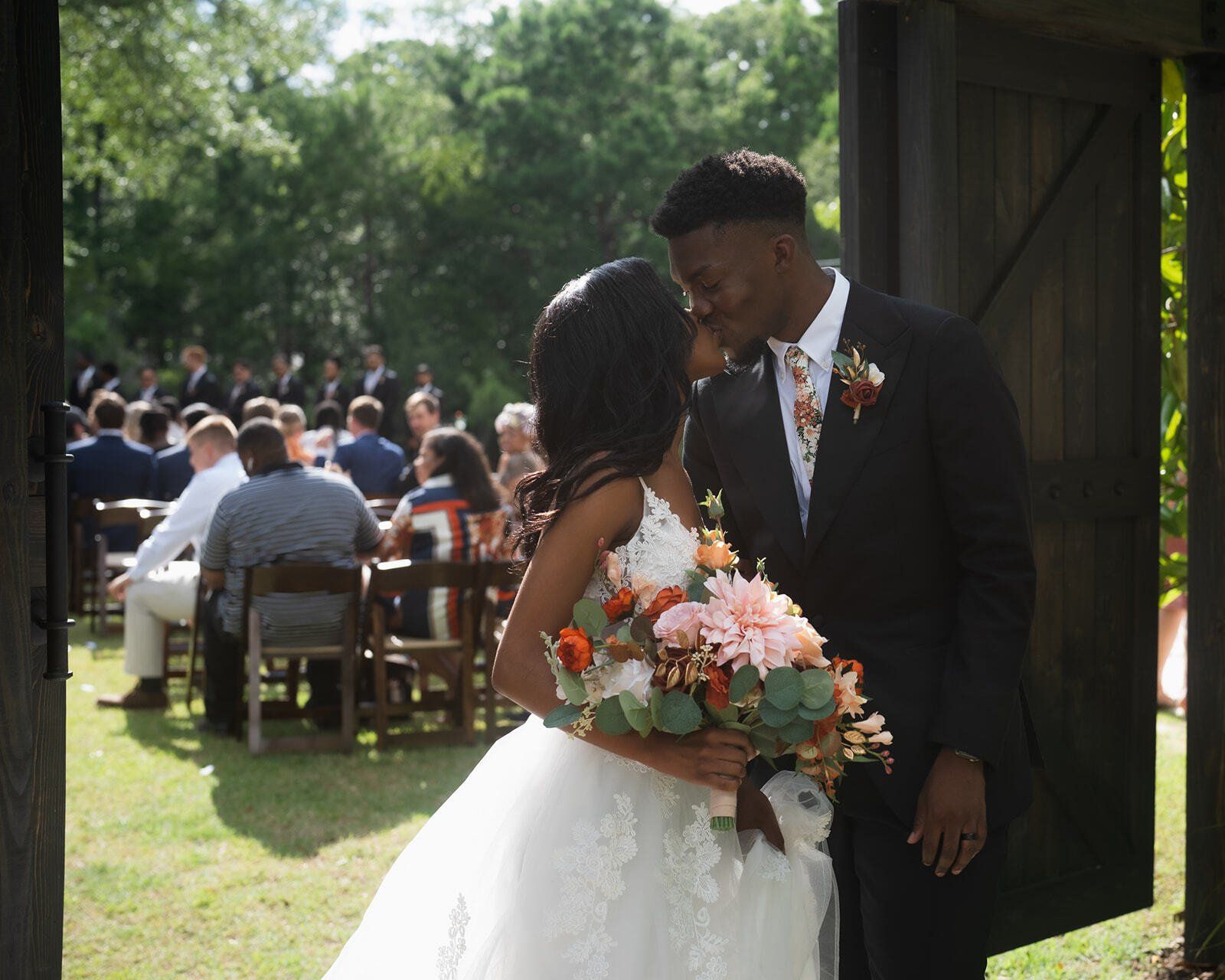 Bride and groom kiss in a sunlit outdoor wedding ceremony, with guests seated behind them.
