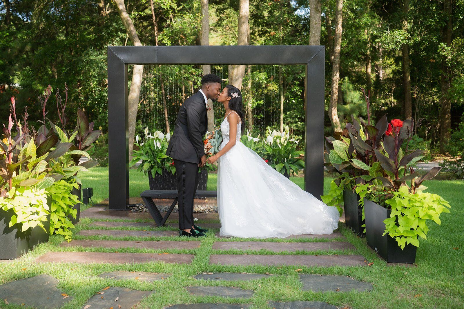 Bride and groom kissing beneath a black garden arch in a lush outdoor wedding setting