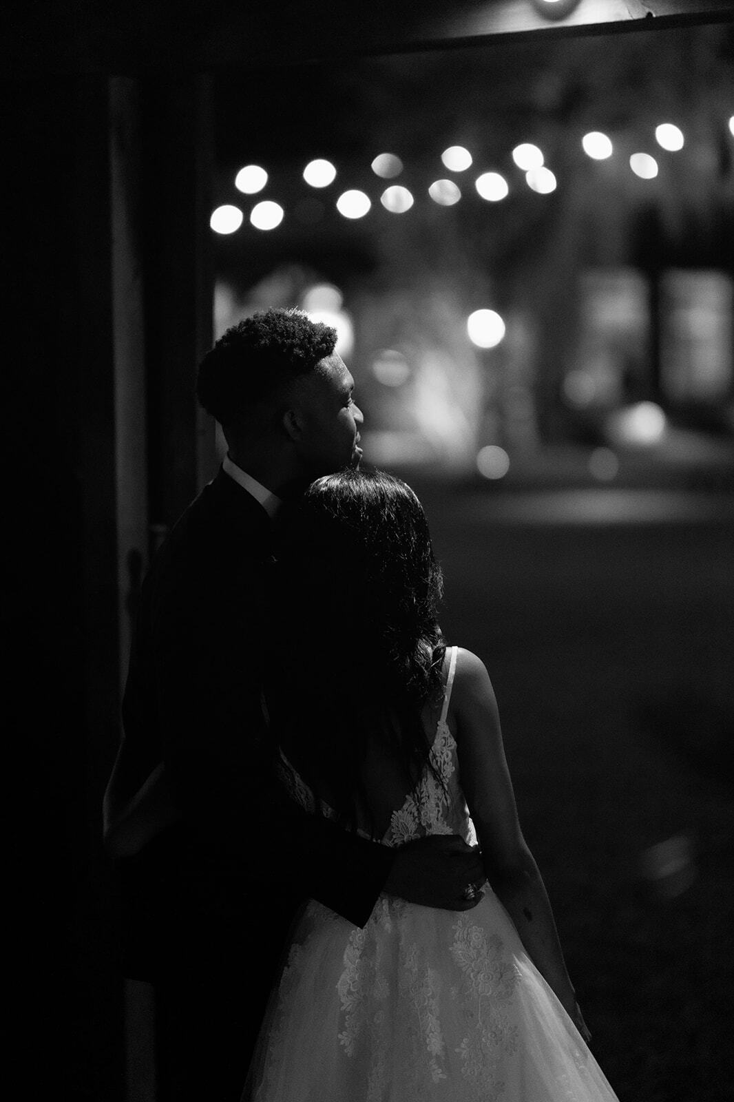Couple embracing under string lights in a dark, black-and-white wedding scene
