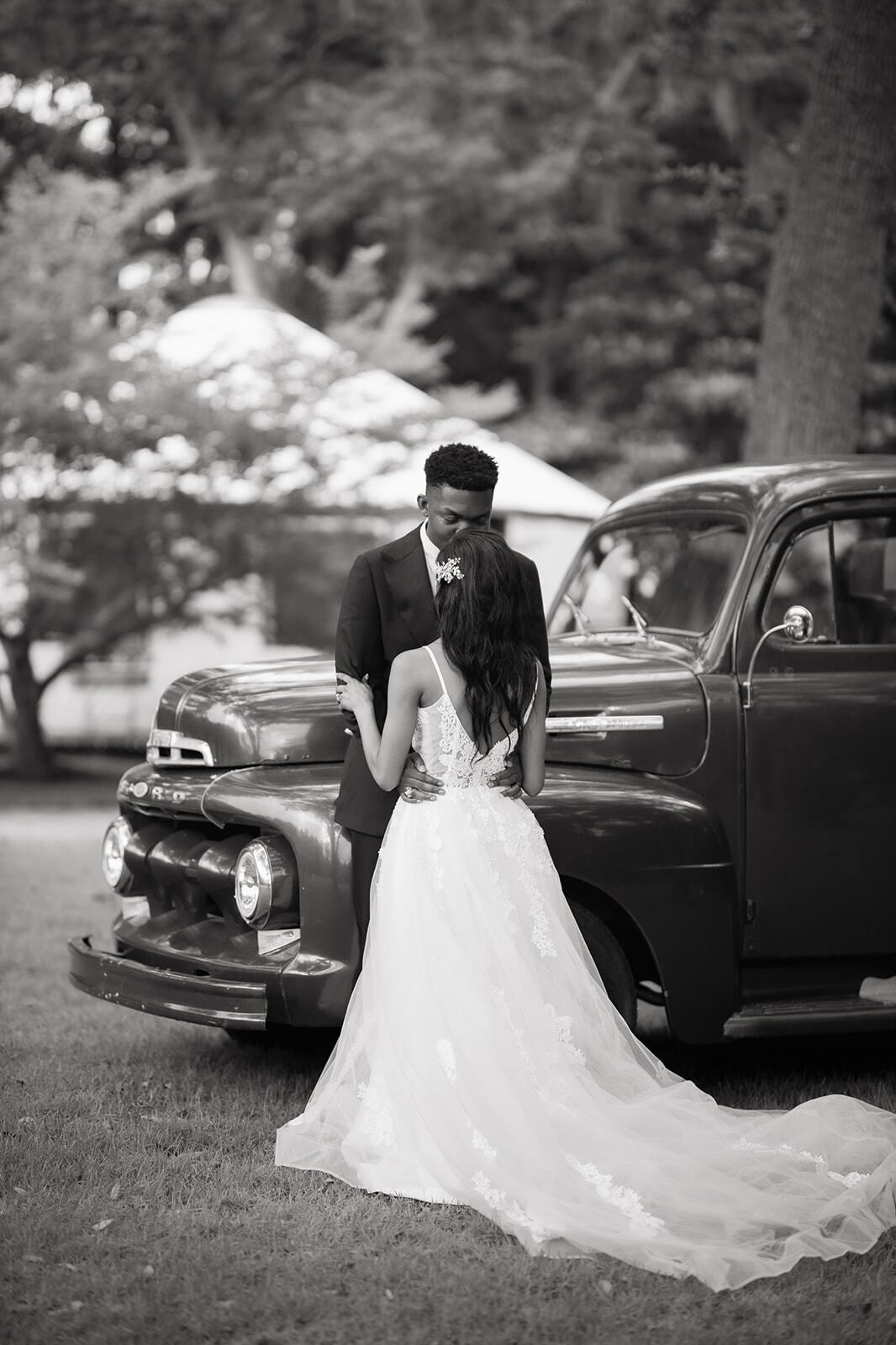 Bride in white gown and groom in dark suit posing beside a vintage car outdoors.