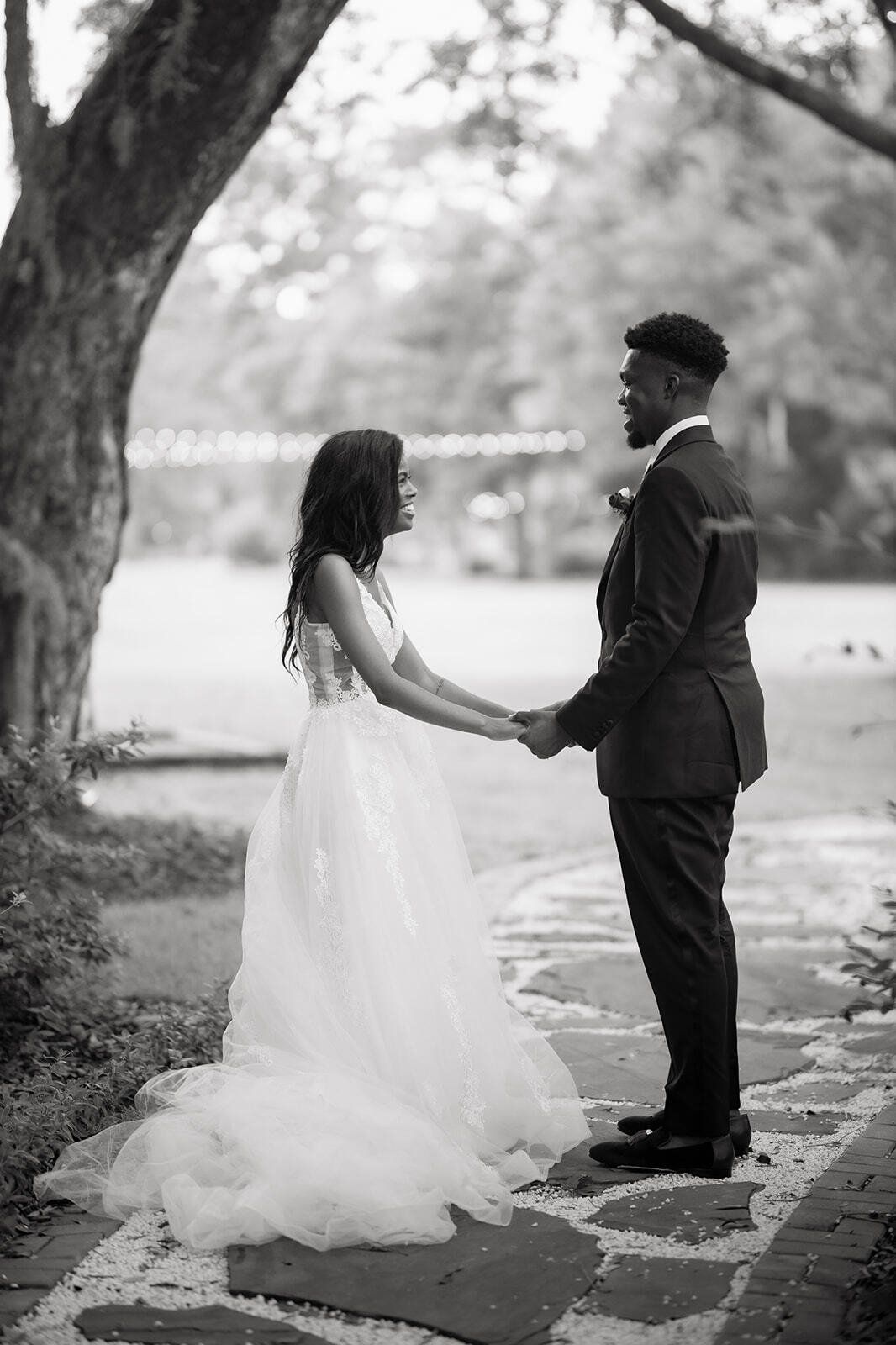 Bride and groom holding hands outdoors under trees, standing on a path in a black-and-white portrait