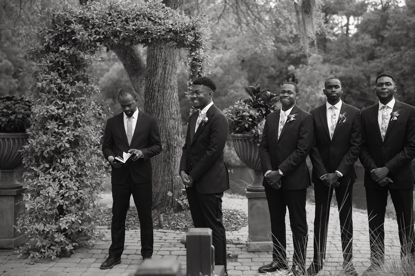 Four men in suits stand under a leafy arch in a garden, posing formally.