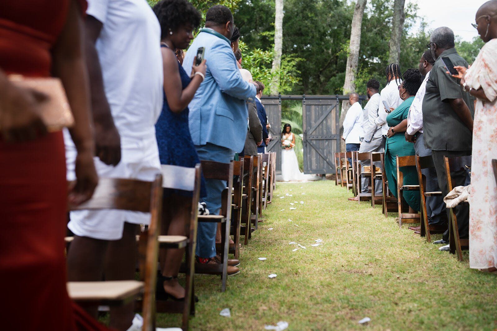 People seated outdoors in rows of wooden chairs along a grassy aisle, facing a central gate