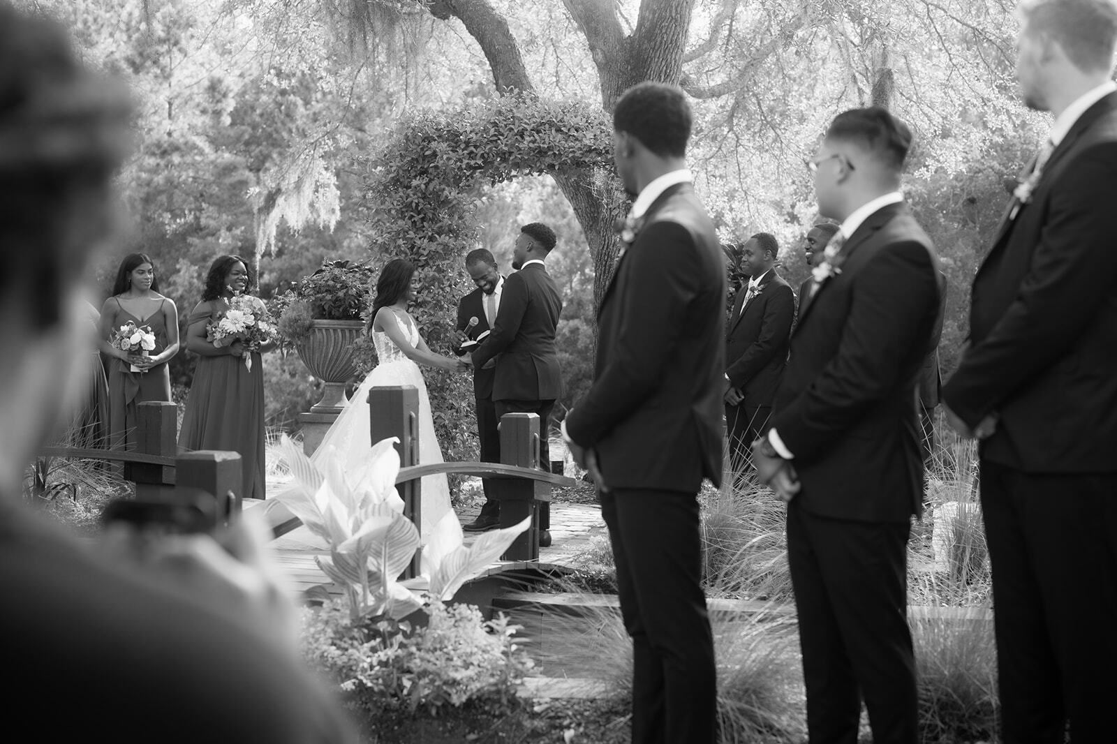 Guests in suits stand at an outdoor wedding ceremony under trees.