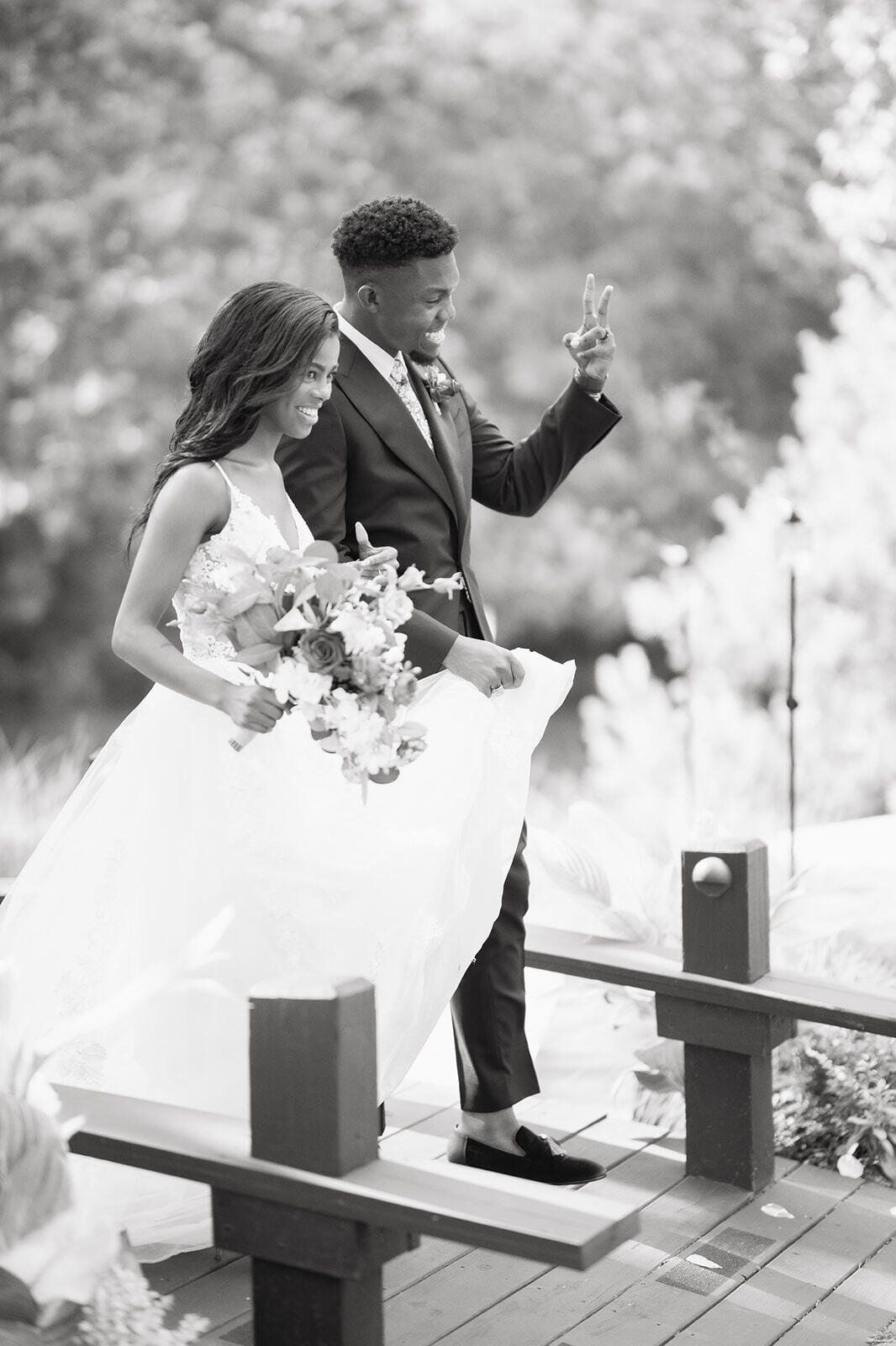 Bride in white gown and groom in dark suit walking outdoors on a wooden bridge, holding flowers and a sparkling wand