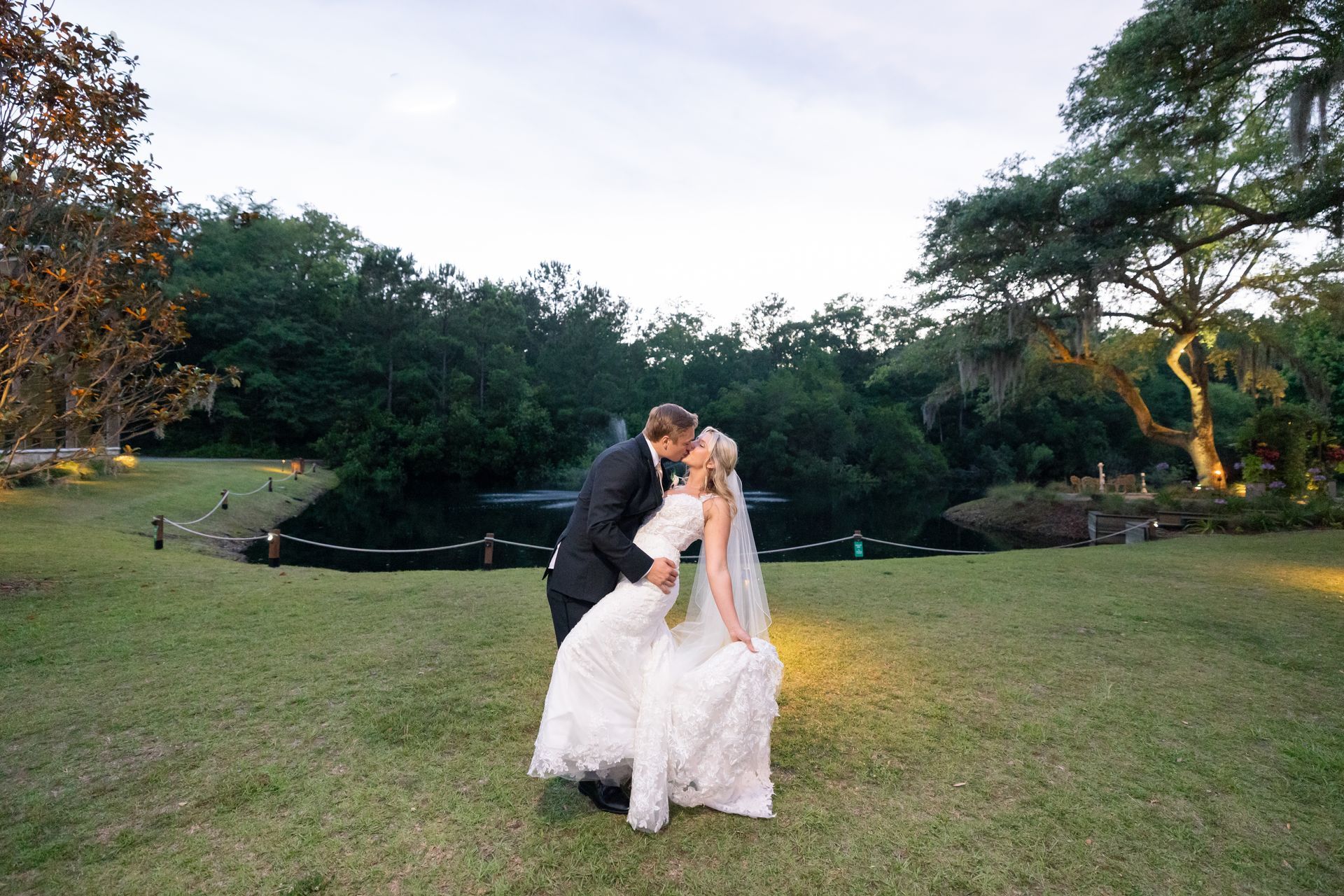 Couple kissing on a grassy lawn at dusk, with trees and a pond in the background.