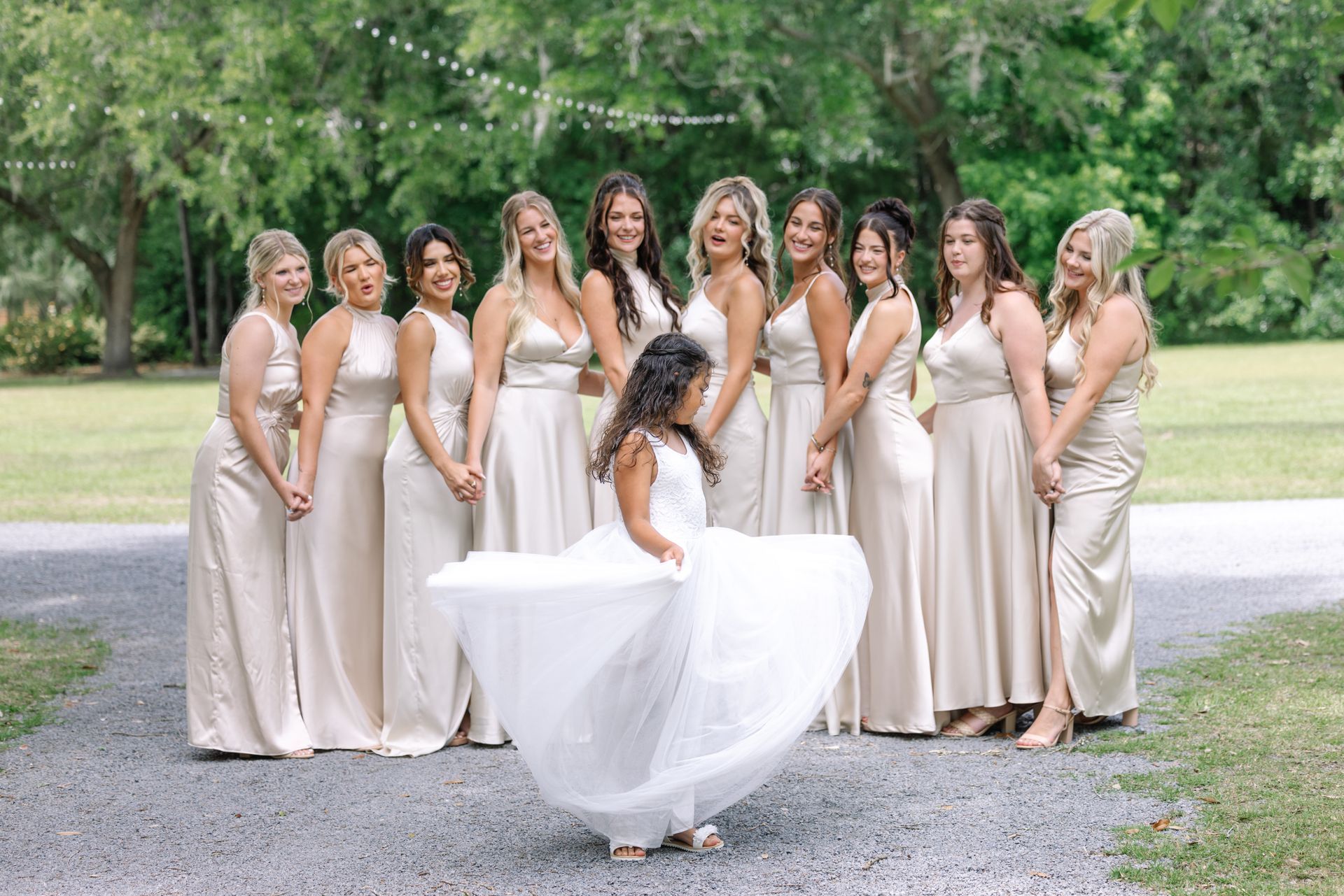 Bridesmaids in champagne dresses pose with a bride holding her gown in a garden setting.