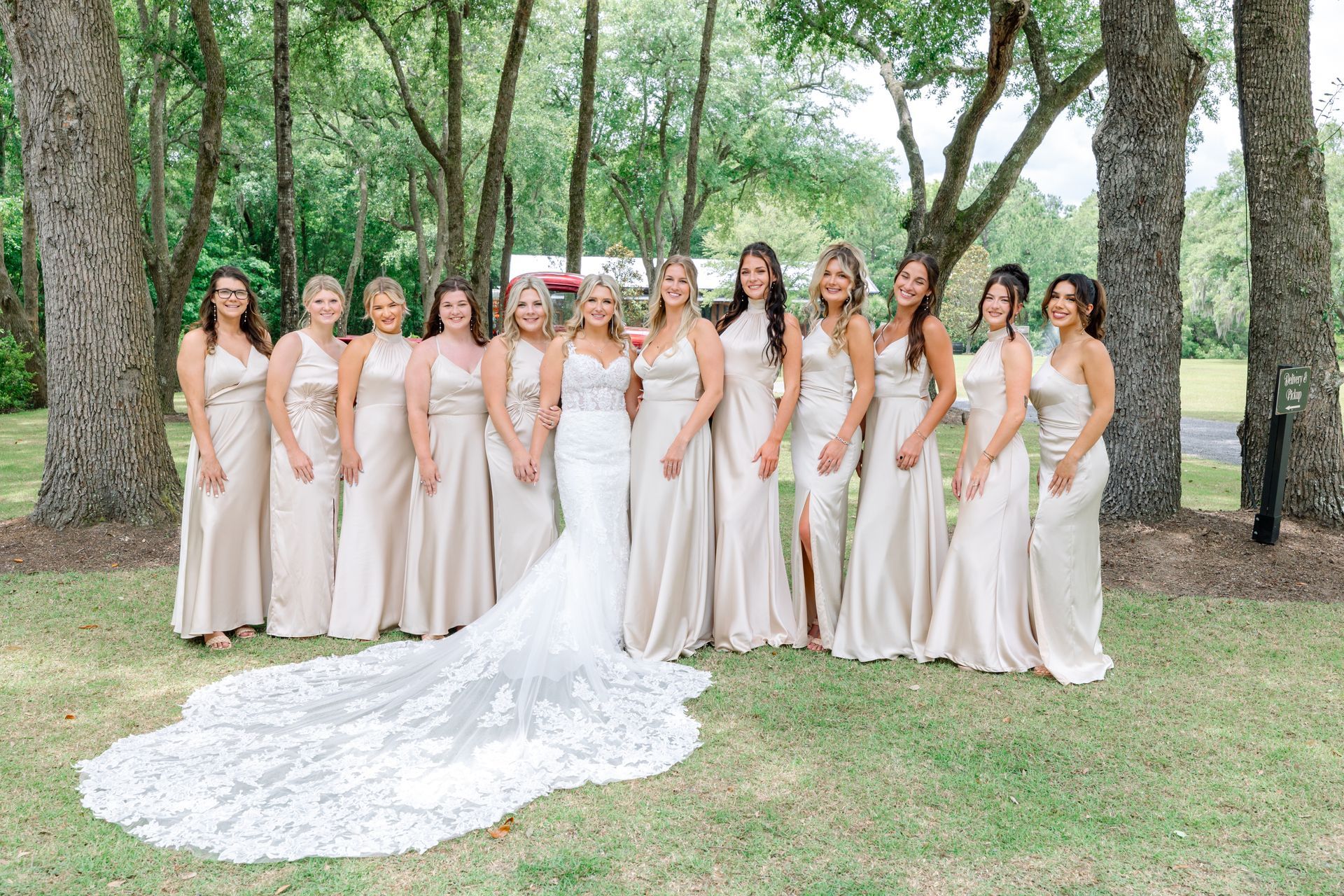Bride and bridesmaids in blush dresses pose under trees, with the bride’s long lace train spread on grass.
