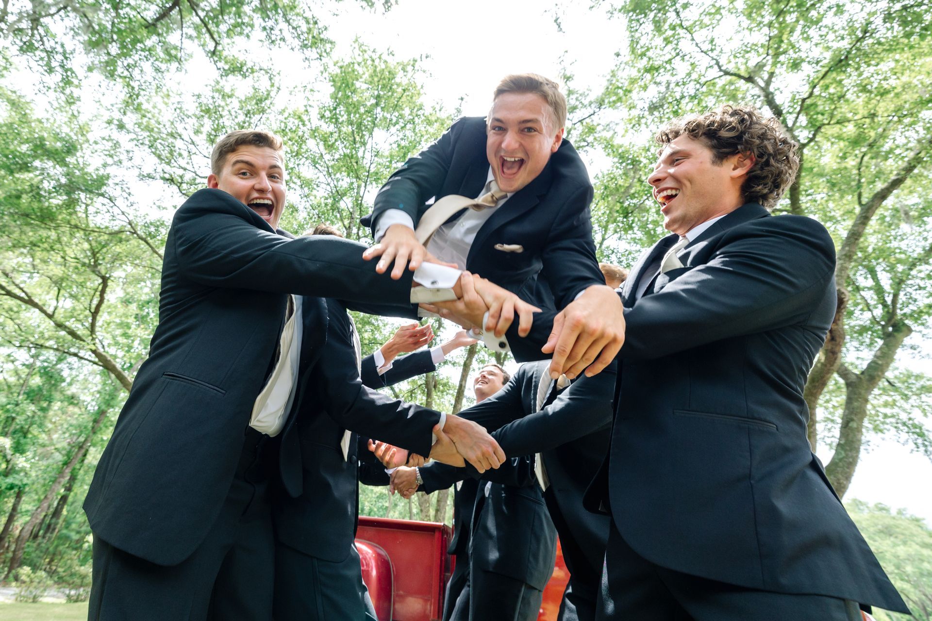 Three men in dark suits stack their hands together and laugh outdoors under trees.