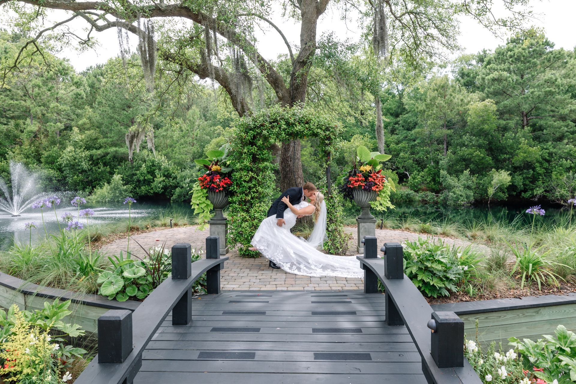 Bride and groom embracing on a garden bridge with red floral arrangements and a fountain nearby