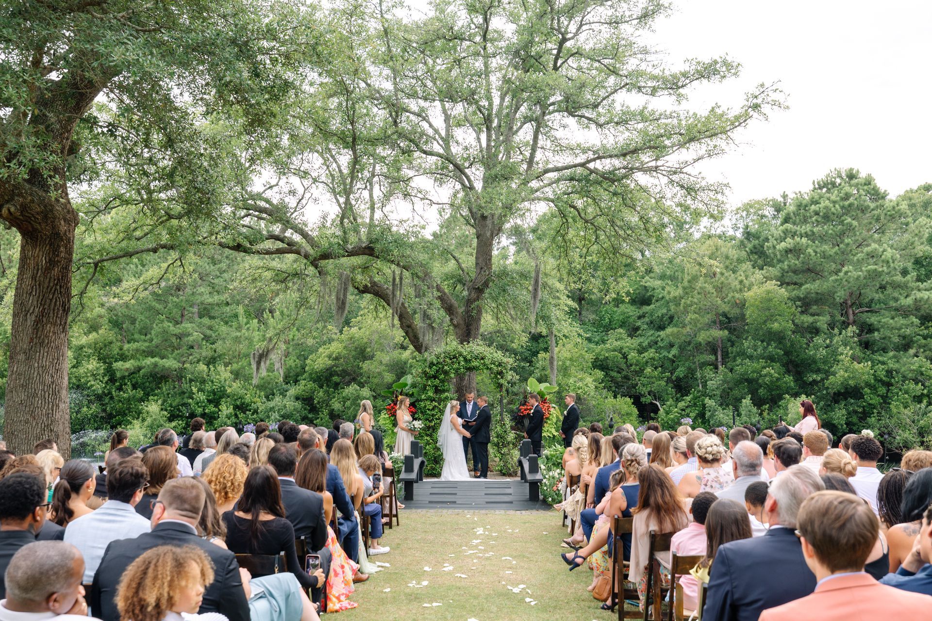 Outdoor wedding ceremony with guests seated on chairs along a tree-lined aisle
