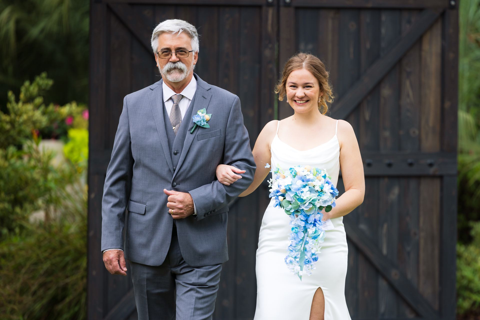 Older man in a gray suit walks beside a bride in a white dress holding a blue-and-white bouquet outdoors