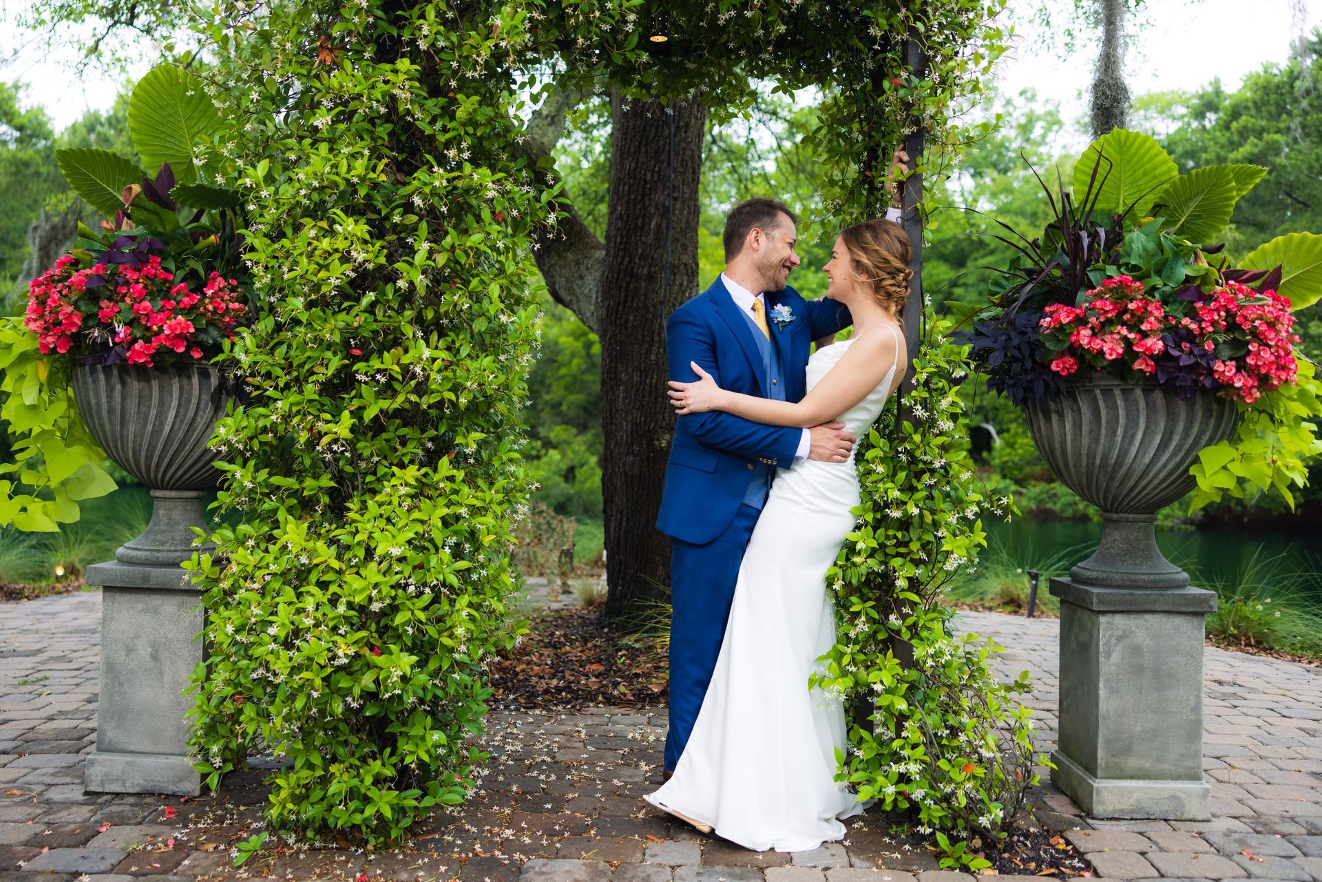 Bride and groom in a garden archway, embracing beneath lush greenery and red flower urns
