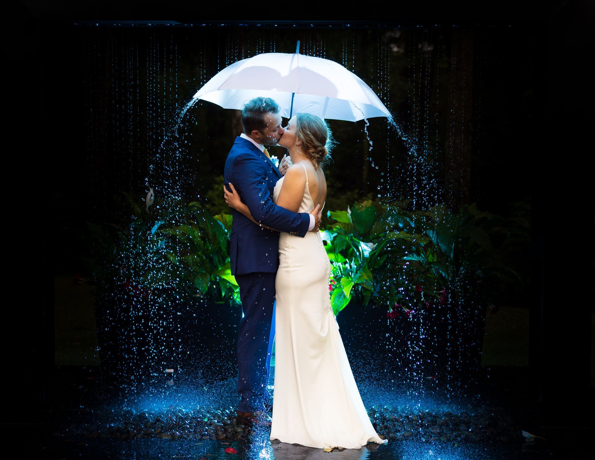 Couple embracing under a white umbrella amid glowing blue rain and greenery