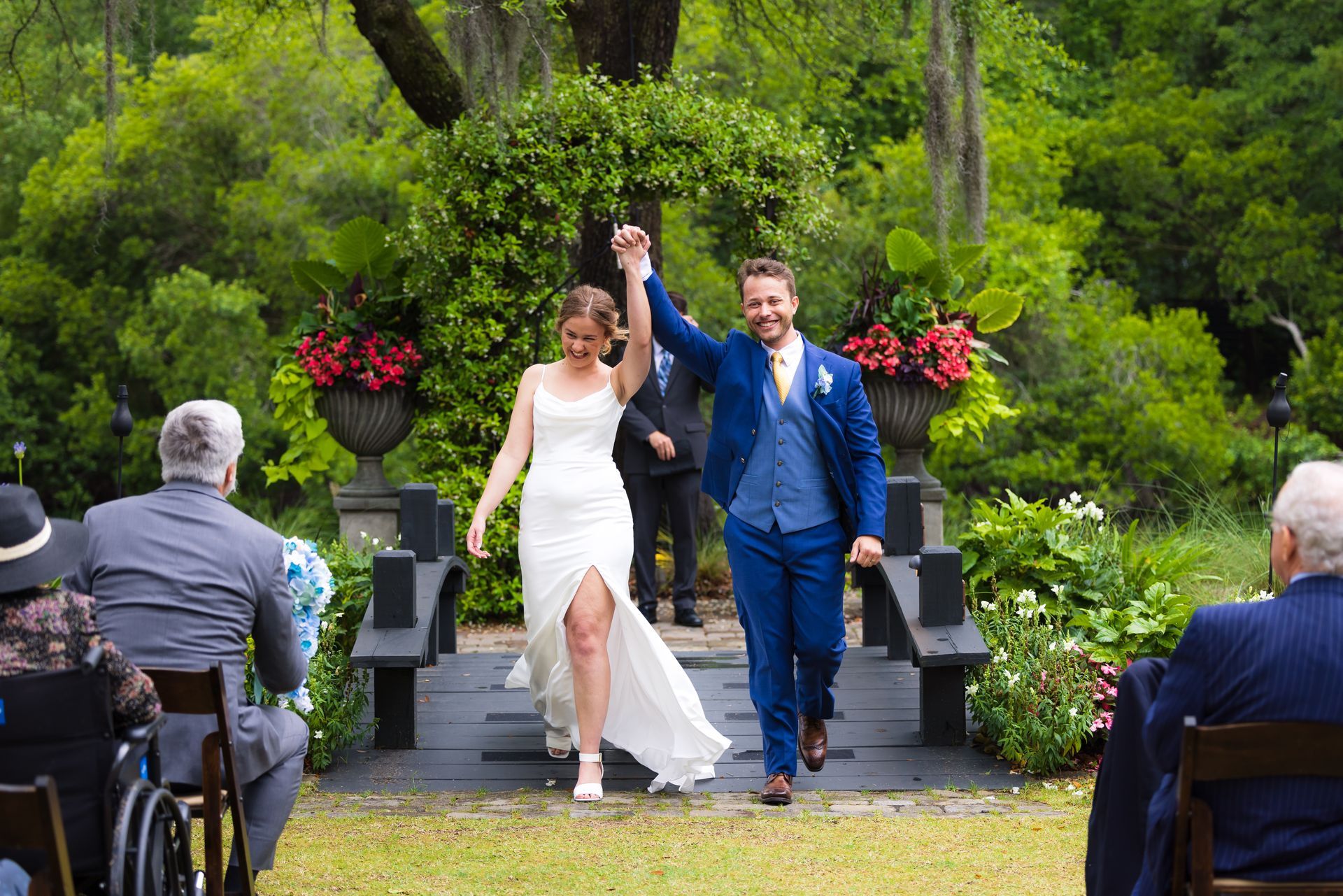 Newlyweds walk down the aisle outdoors, holding hands and smiling, with guests seated on either side.