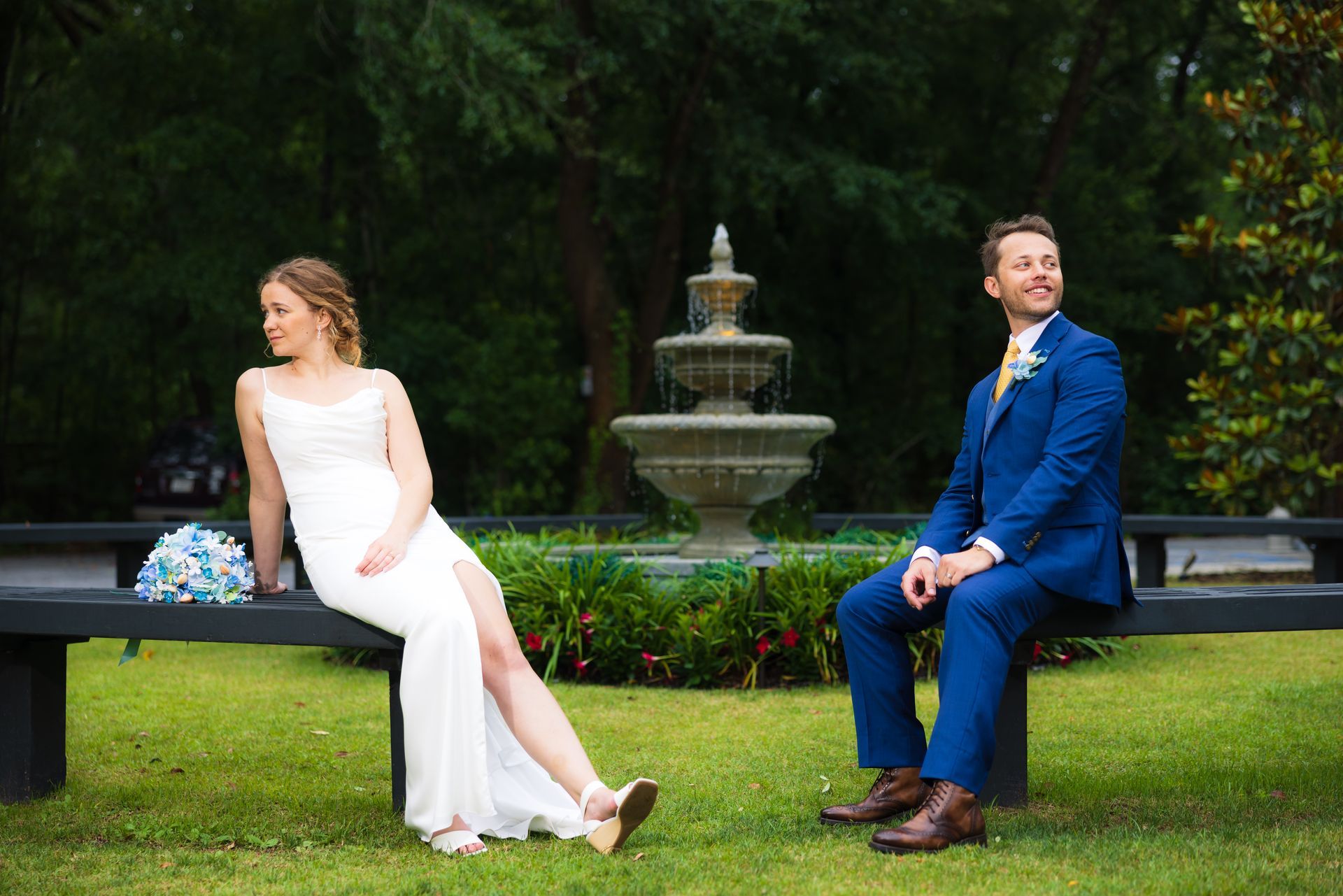 Couple in wedding attire seated apart on a lawn with a fountain in the background.