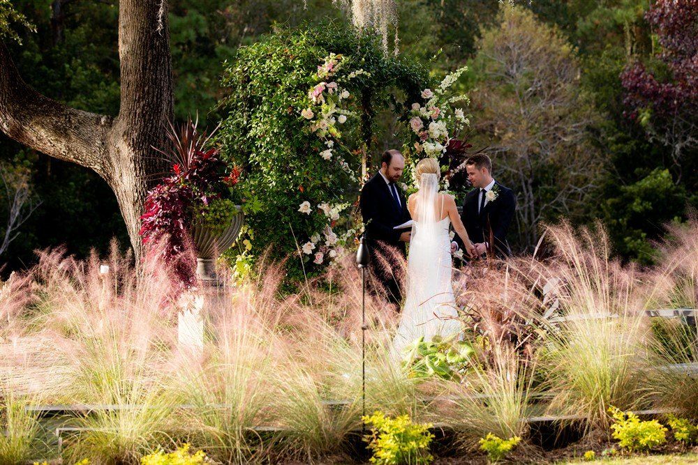 Bride and groom stand beneath a floral arch in a garden among tall grasses and trees.