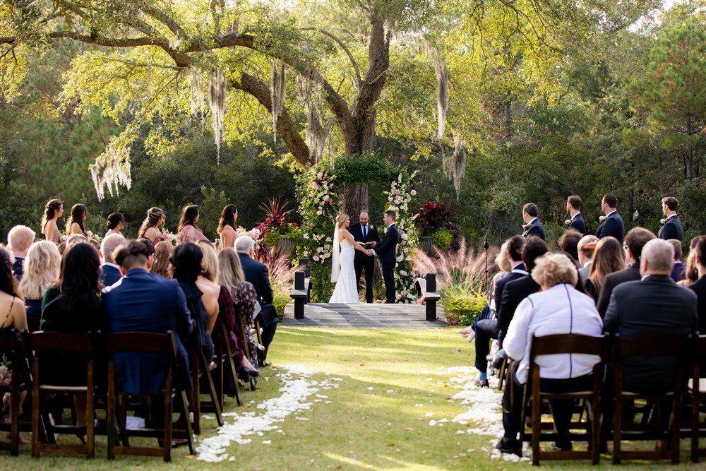 Outdoor wedding ceremony with bride and groom under trees, guests seated on both sides of the aisle