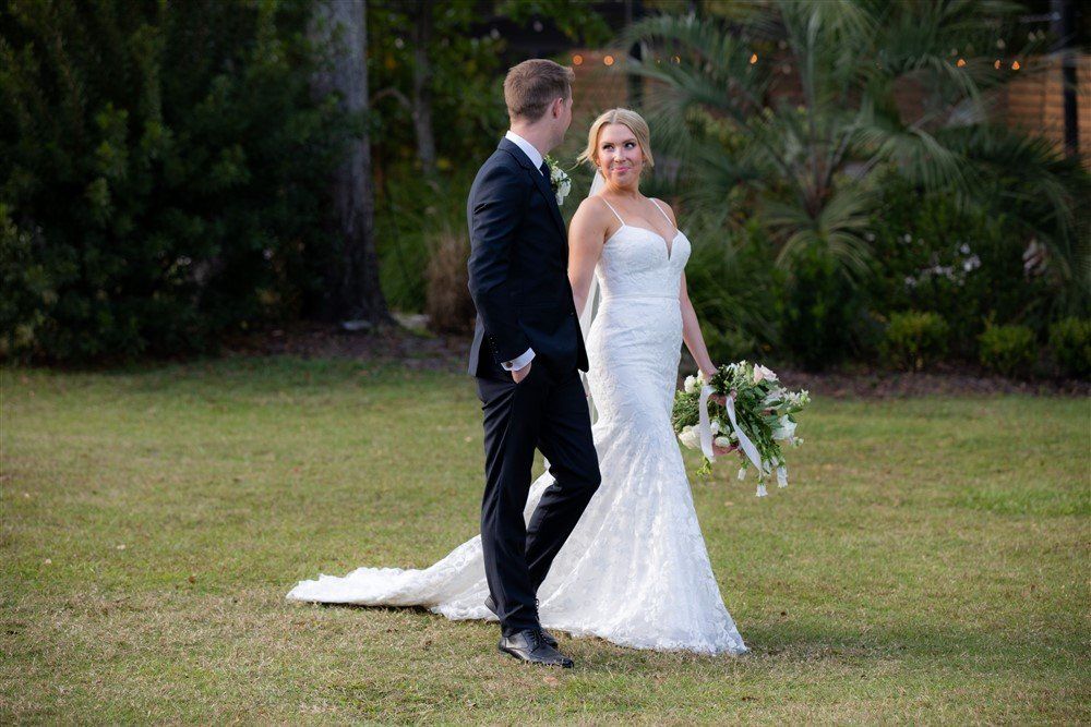 Bride and groom pose outdoors on a lawn, the bride holding a bouquet and wearing a white gown.