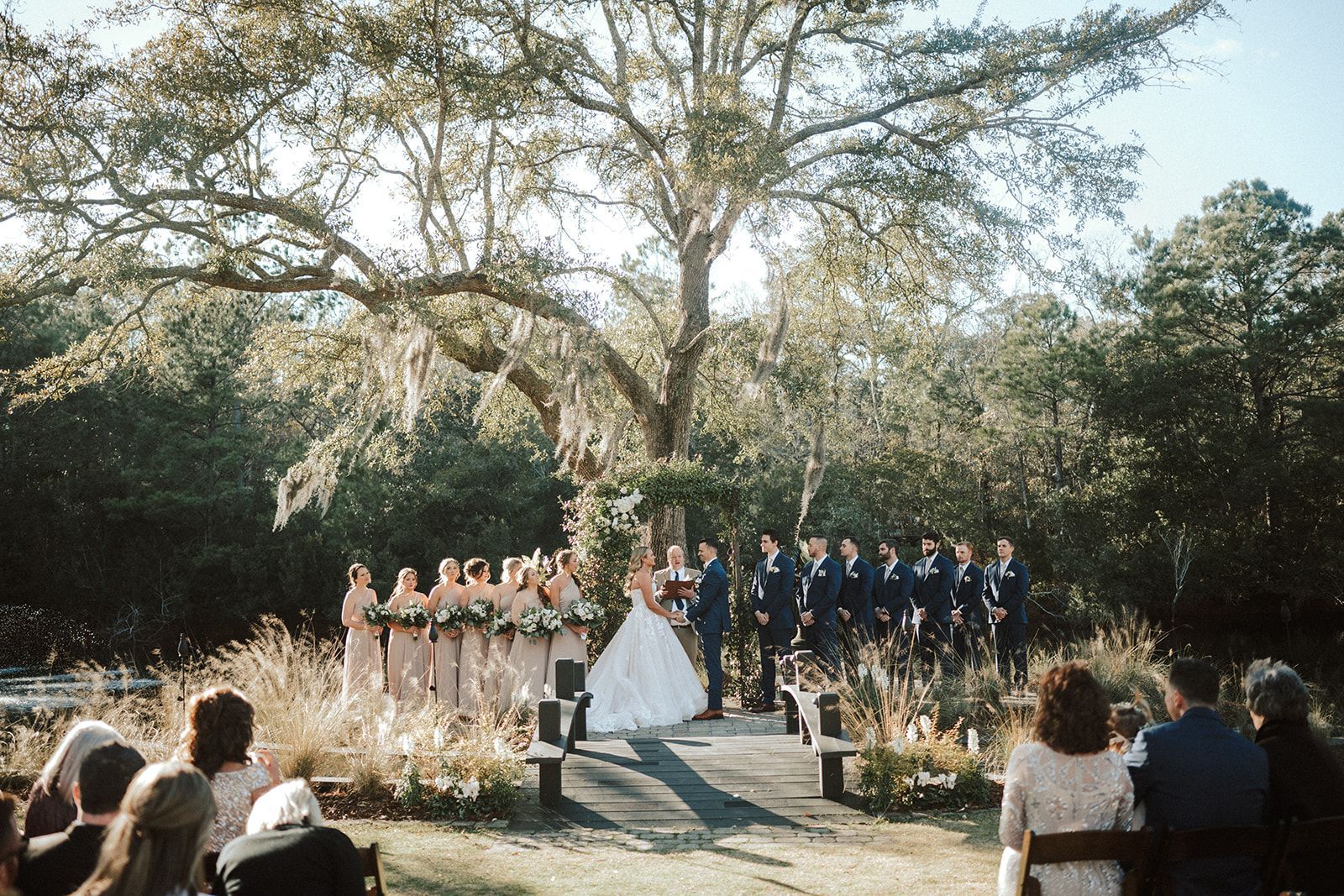 Outdoor wedding ceremony under a large tree, with bride, groom, and bridal party facing guests on wooden chairs.