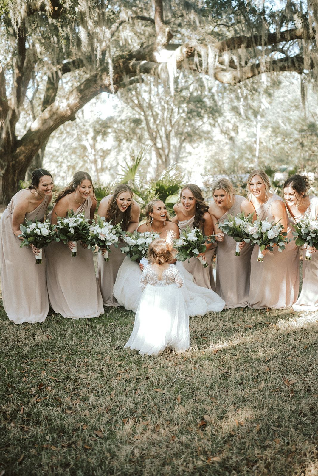 Bride with bridesmaids holding bouquets in a grassy outdoor wedding setting under hanging moss.