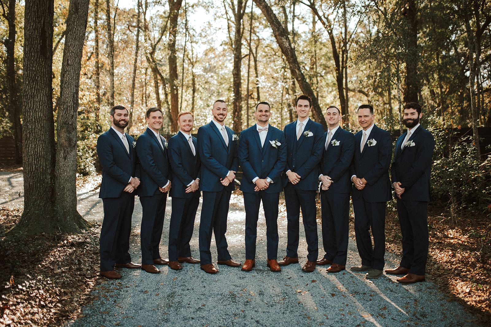 Seven men in dark suits standing together on a gravel path in a wooded outdoor setting.