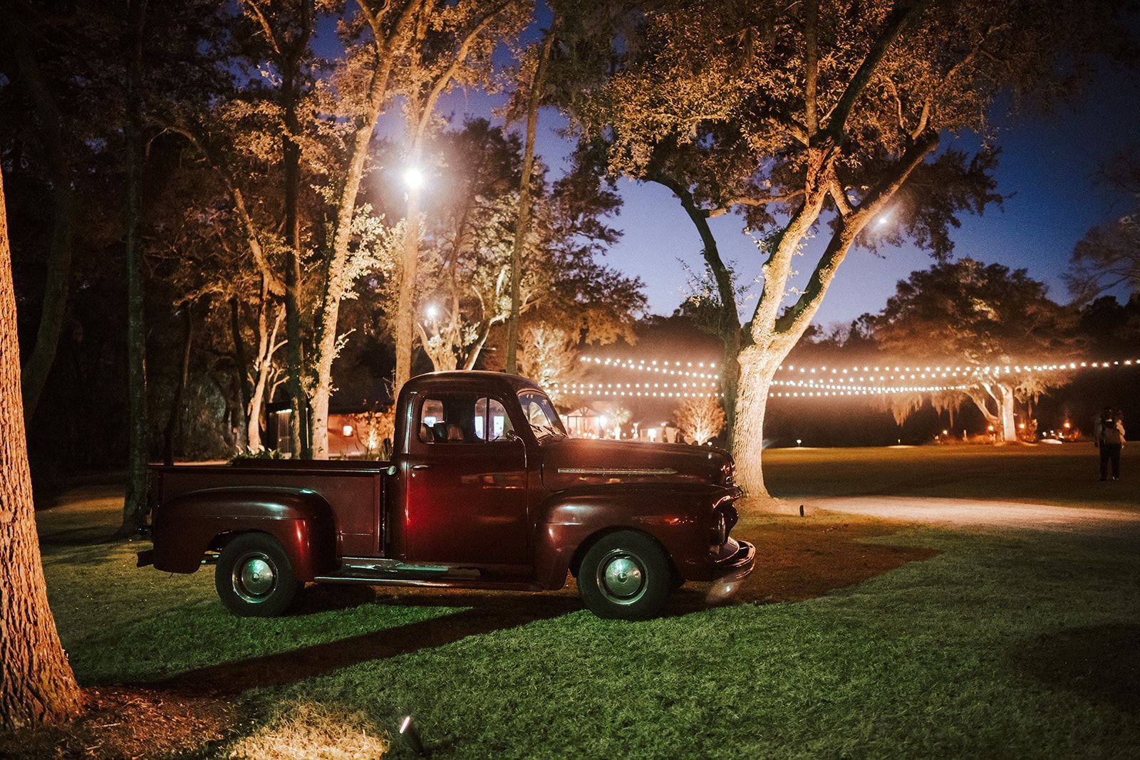 Red pickup truck parked on grass under trees at night with warm lights in the background