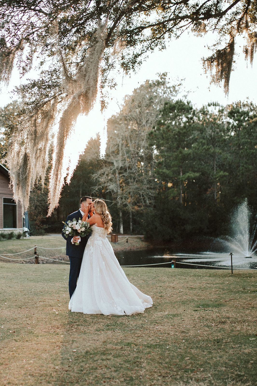 Bride and groom standing outdoors under hanging moss, embracing in a grassy garden at sunset